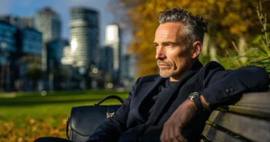 En medelålders man sitter på en parkbänk med en modern stadssilhuett i bakgrunden. Han bär en stilren kavaj och har en professionell väska bredvid sig. Midcent. Kategori: Samhälle.