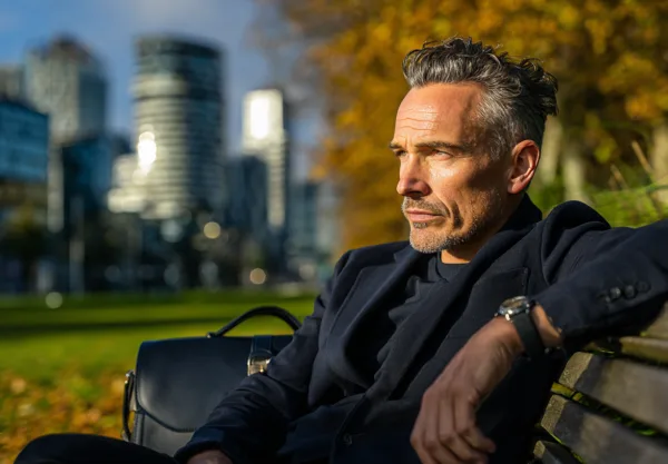 En medelålders man sitter på en parkbänk med en modern stadssilhuett i bakgrunden. Han bär en stilren kavaj och har en professionell väska bredvid sig. Midcent. Kategori: Samhälle.