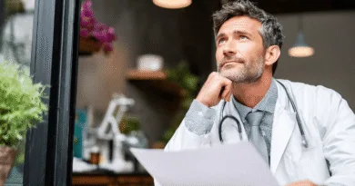 En medelålders man i vit läkarrock sitter fundersamt vid ett bord, omgiven av gröna växter och instrument. Han håller ett papper och bär ett stetoskop. Midcent: Hälsa.