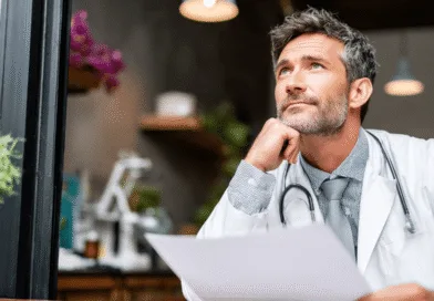 En medelålders man i vit läkarrock sitter fundersamt vid ett bord, omgiven av gröna växter och instrument. Han håller ett papper och bär ett stetoskop. Midcent: Hälsa.