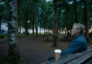 Midcent: En medelålders man sitter på en parkbänk omgiven av grönska i stadsmiljö, med en kaffemugg bredvid sig, vilket skapar en avkopplande atmosfär i kvällsljus. Kategori: Samhälle.