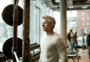 En medelålders man står i ett modernt gym med vikter i fokus, vilket speglar styrka och hängivenhet inom fitness. Ljus interiör och engagerad atmosfär. Midcent - Hälsa.