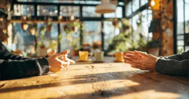 Midcent: Två personer samtalar vid ett träbord i en solig kafémiljö, med gröna växter i bakgrunden. Perfekt söndagsfika i mysig atmosfär. Underhållning.