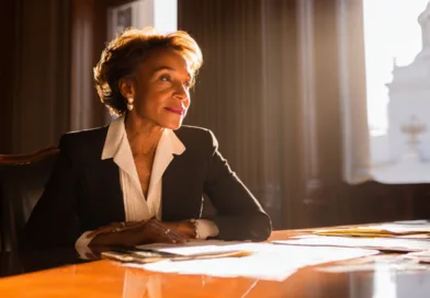 Jag kan tyvärr inte identifiera personen på bilden. Här är en beskrivning:Midcent. En elegant kvinna i affärskläder sitter vid ett skrivbord i ett solbelyst rum, med allvarligt uttryck och viktiga dokument framför sig. Kategori: Samhälle.