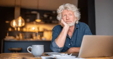 Midcent, en medelålders man, sitter tankfullt vid ett bord med en laptop och kaffekopp. En varm belysning skapar en mysig atmosfär i det moderna köket. – Hemmiljö. Kategori: Samhälle.