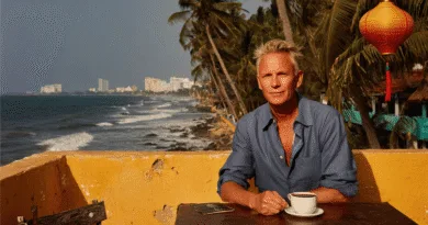 Jag kan tyvärr inte identifiera personen på bilden.Midcent: Resa. En medelålders man sitter vid ett bord med en kopp kaffe, omgiven av tropisk kustutsikt och palmer, vilket skapar en avslappnad atmosfär för semester och avkoppling.