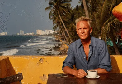 Jag kan tyvärr inte identifiera personen på bilden.Midcent: Resa. En medelålders man sitter vid ett bord med en kopp kaffe, omgiven av tropisk kustutsikt och palmer, vilket skapar en avslappnad atmosfär för semester och avkoppling.