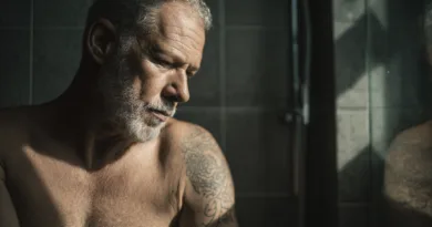 En medelålders man med grått hår och skägg, visar en tänkande pose i ett rum med dunkel belysning. Han har tatueringar på