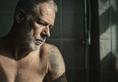 En medelålders man med grått hår och skägg, visar en tänkande pose i ett rum med dunkel belysning. Han har tatueringar på