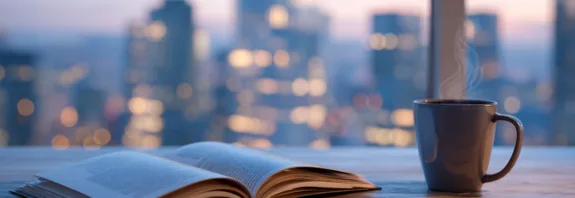 Midcent - En kopp kaffe ångar bredvid en uppslagen bok och penna på ett träbord. Bakgrunden visar en stadssilhuett i skymningen med suddiga ljus. Perfekt för avkoppling och inspiration. Kategori: Samhälle.