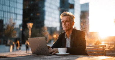 Midcent: En medelålders man arbetar utomhus med en laptop, omgiven av moderna byggnader och solnedgångens sken. Han verkar fokuserad och reflekterande, med en kopp kaffe vid sin sida. Kategori: Samhälle.