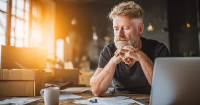 Midcent: En medelålders man sitter vid ett skrivbord med dator och papper, djup i tankar. Solen strömmar in genom fönstren och skapar en varm atmosfär. Kategorin: Ekonomi.