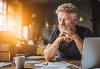 Midcent: En medelålders man sitter vid ett skrivbord med dator och papper, djup i tankar. Solen strömmar in genom fönstren och skapar en varm atmosfär. Kategorin: Ekonomi.