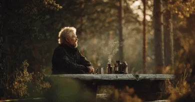 Midcent: En medelålders man njuter av en lugn stund vid en picknickbänk i skogen, omgiven av termosar och naturens rogivande miljö. Solen lyser genom träden och skapar en fridfull atmosfär. Kategori: Resa.