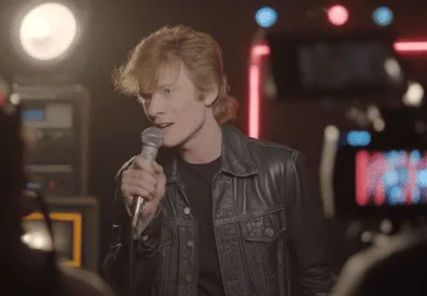 Midcent: En medelålders man sjunger passionerat i en inspelningsstudio, omgiven av ljus och kamerautrustning. Hans rockiga stil kompletteras av en läderjacka, vilket skapar en stämningsfull atmosfär. Underhållning.