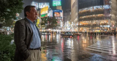 Midcent: En medelålders man står i regnet vid ett livligt stadslandskap med upplysta skyltar och reflekterande våta gator. Stadens puls och nattliv skapar en dynamisk atmosfär. Kategori: Resa.