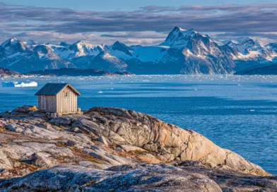 Midcent Resa: Upptäck storslagen skönhet med en liten stuga på klippiga kuster med snötäckta berg i bakgrunden. Fantastiska vyer över isberg och det klarblå havet. Perfekt för naturälskare och äventyrsresenärer.