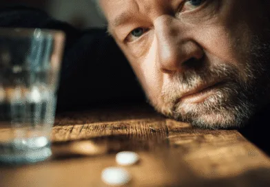 En medelålders man ser tankfull ut vid ett bord med ett glas vatten och två tabletter. Bilden fångar en känsla av reflektion och eftertanke. Midcent. Hälsa.