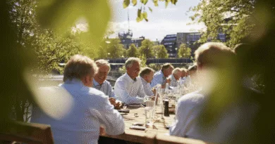 Midcent - En medelålders man deltar i en utomhusaffärslunch med kollegor på en solig dag. Mötet sker runt ett stort träbord, omgiven av grönska och stadsvyer. Ekonomi.