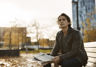 Jag kan tyvärr inte identifiera personen på bilden. Här är en beskrivning:Midcent: En medelålders man sitter på en bänk i urban miljö med en tidning i handen. Höstlöv runtomkring skapar en rofylld atmosfär. Samhälle.