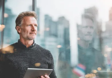 En medelålders man står vid ett fönster i en stadsmiljö, hållande en surfplatta. Bilden förmedlar en känsla av reflektion och digital anslutning. Midcent. Kategori: Teknik.