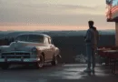 Midcent: En ung man i jeansjacka står bredvid en klassisk bil utanför ett retro-inspirerat kafé vid solnedgången. Lugnt landskap i bakgrunden, vilket skapar en nostalgisk känsla. Kategori: Resa.