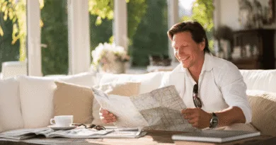 En medelålders man ler och granskar en karta i ett soligt vardagsrum. Han sitter avslappnad med en kaffekopp på ett soffbord. En atmosfär av avkoppling och planering genomsyrar bilden.Midcent Resa.